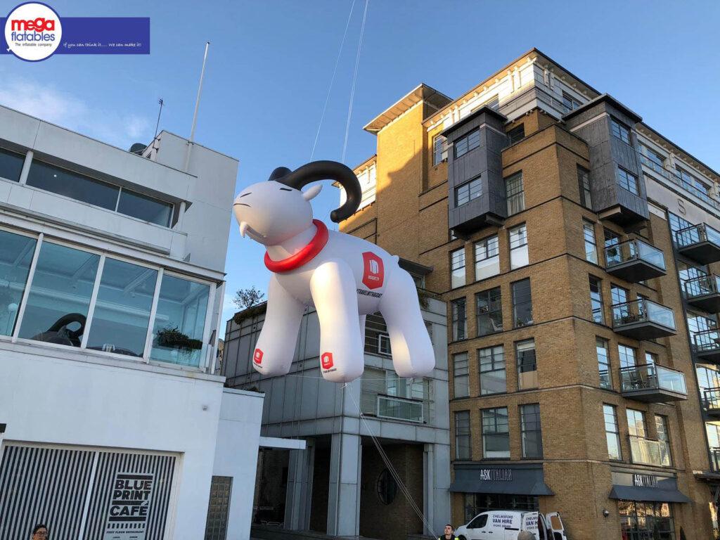 Large inflatable white goat with black horns and a red collar, branded with “D” logos, displayed outdoors near modern buildings and the Blue Print Café.