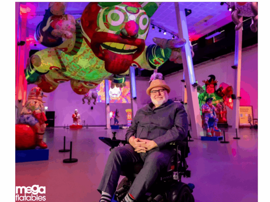 A man sits smiling in front of a colourful exhibition filled with large inflatable sculptures suspended from the ceiling. The space glows with vibrant lighting, highlighting the playful, detailed artwork. These inflatables were created by Mega Inflatables in collaboration with digital artist Jason Wilsher-Mills, transforming his imaginative digital pieces into large-scale 3D installations.