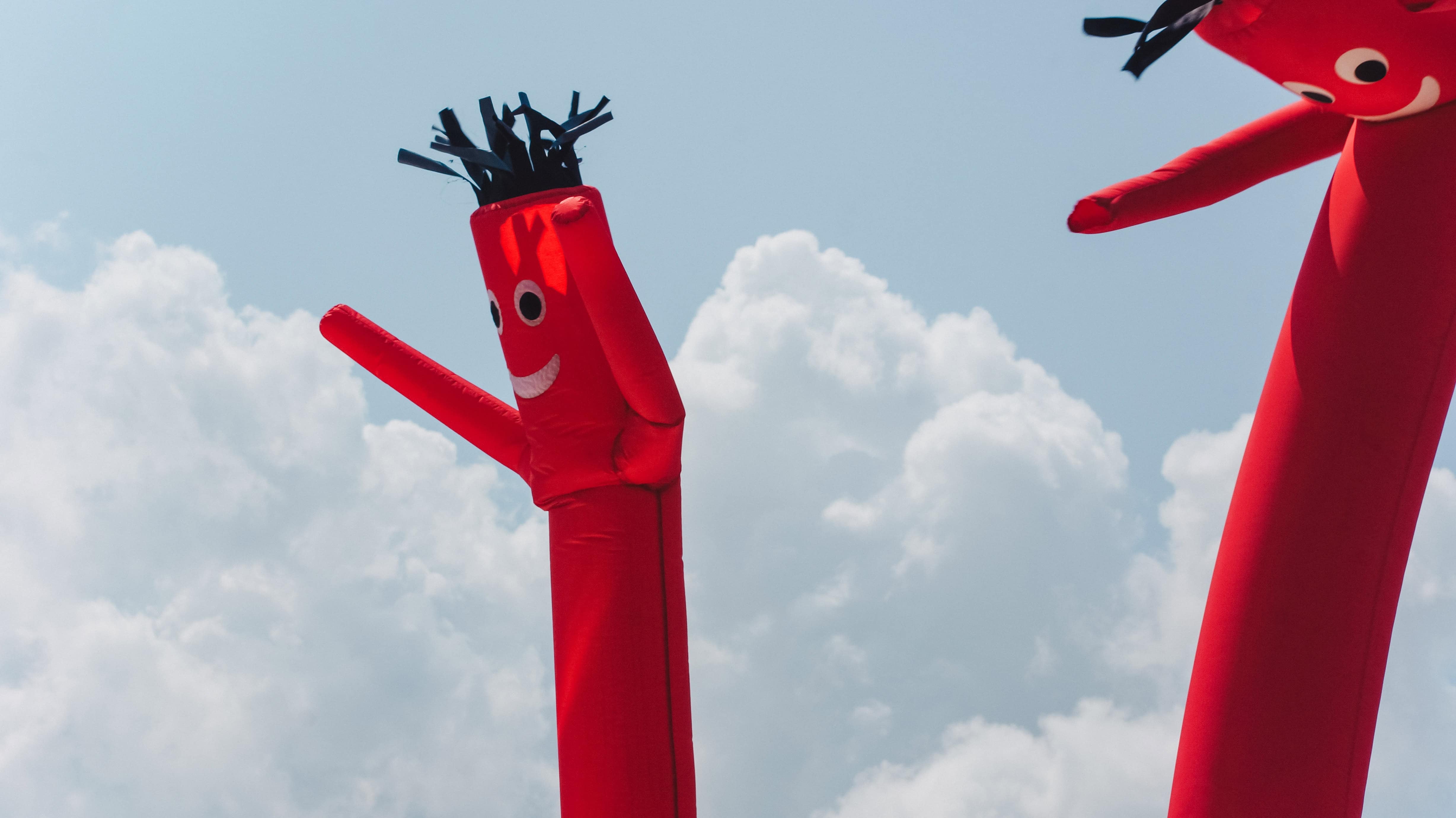 large red inflatable dancing tube man with a smiling face and black hair waving in the wind against a cloudy sky